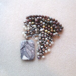 May include: A collection of beads in various colors and sizes, including brown, white, and gray. The beads are arranged in a pile on a white surface. A rectangular gray and white stone pendant is also included.