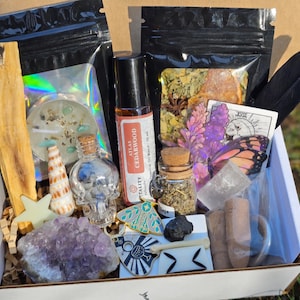 May include: A gift box containing a palo santo stick, a skull-shaped glass bottle, a cedarwood essential oil roller, a small glass jar with herbs, an amethyst cluster, a seashell, a black feather, incense cones, and various small decorative charms.