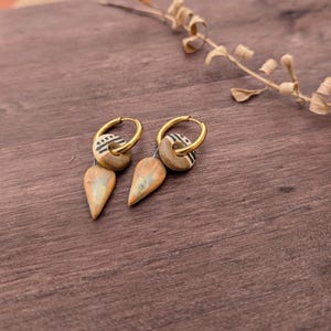 Può includere: Un paio di orecchini a cerchio color oro con ciondoli pendenti. Ogni ciondolo è composto da una piccola perla rotonda color marrone chiaro con motivi a linee nere e un ciondolo in ceramica smaltata a forma di goccia color marrone chiaro e verde.