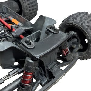 May include: A black 3D-printed shock tower brace installed on a remote-controlled vehicle. The component features two mounting points with black plastic posts, positioned above red coil-over shock absorbers and large, knobby black rubber tires.