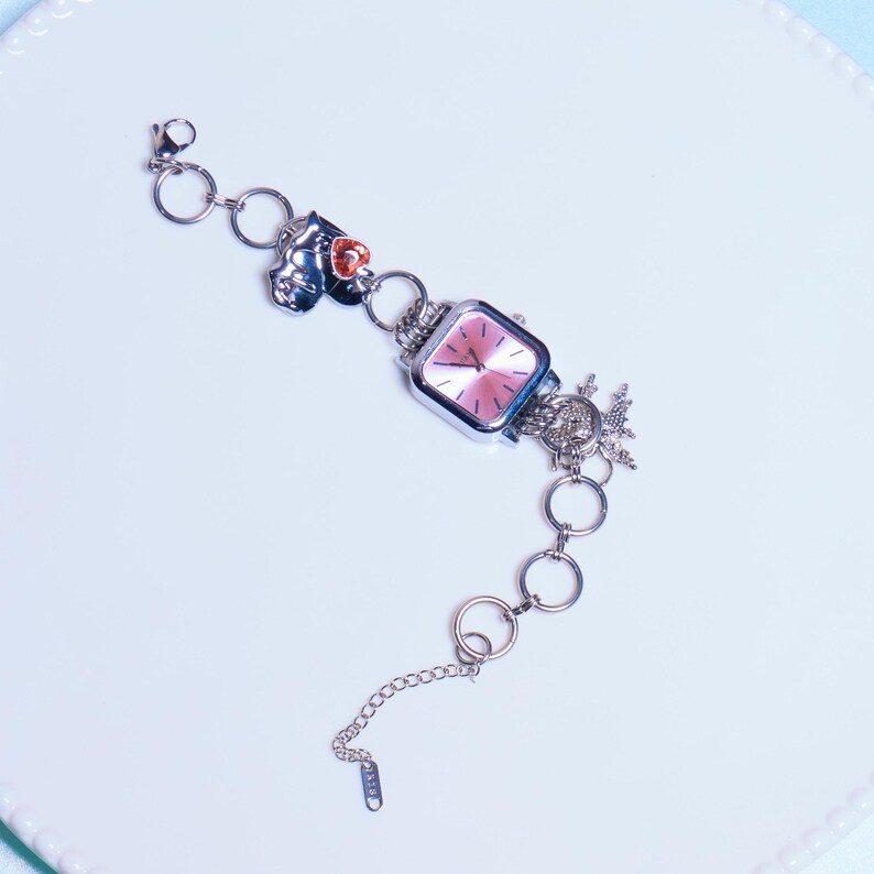 Op de afbeelding: Een zilveren armband met bedels, waaronder een horloge met een roze wijzerplaat, een zilveren hartbedel en een zilveren bloemenbedel.