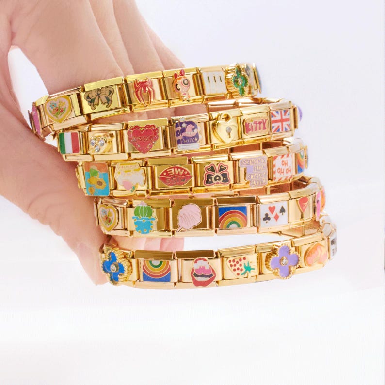 May include: Gold charm bracelets with colourful enamel charms. The charms feature various designs, including hearts, flags, flowers, and rainbows. The bracelets are stacked, showcasing the variety of charms and the gold metal.