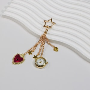 May include: Gold-toned charm with a star-shaped clasp, chains, and dangling charms. Charms include a red heart, stars, a small watch, and a heart. The watch has a white face with black numbers and hands. The charms are on a white background.