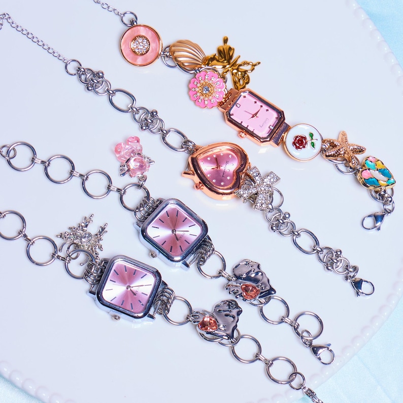 Op de afbeelding: Vier zilveren armbandjes met roze wijzerplaten en verschillende bedels, waaronder een hart, een vlinder, een bloem, een ster en een schelp.