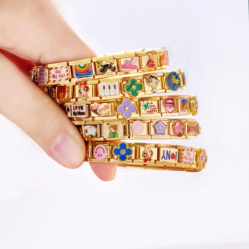 May include: Gold charm bracelets with colourful enamel charms. Each bracelet features various charms, including a rainbow, piano keys, flowers, and the text "NY". The bracelets are held in a hand.