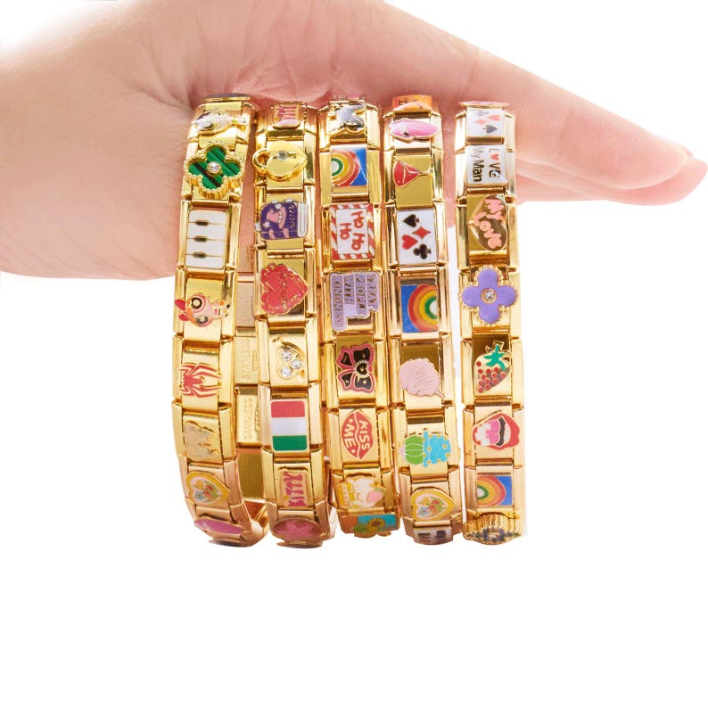 May include: Several gold-coloured charm bracelets, each made of rectangular links decorated with colourful enamel charms. The charms display diverse designs, including flowers, hearts, and rainbows. The bracelets are held against a white backdrop.
