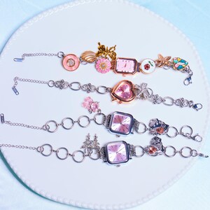 Op de afbeelding: Vier zilveren armbandjes met roze wijzerplaten en verschillende bedels, waaronder bloemen, vlinders en harten. De armbandjes liggen op een witte achtergrond.