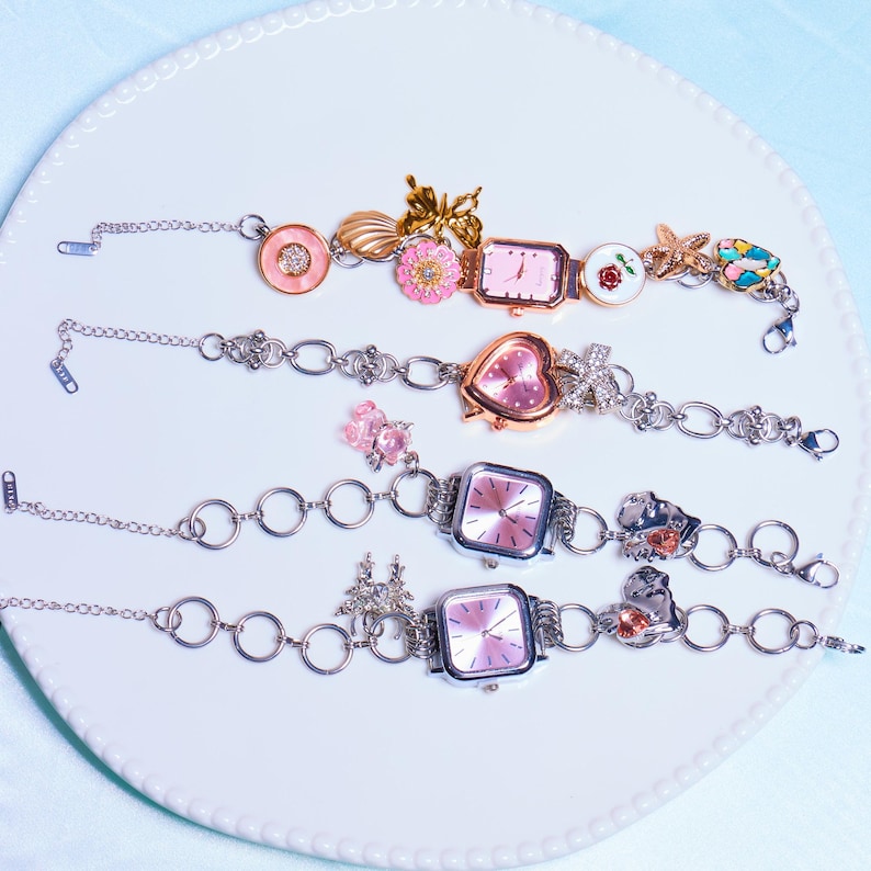 Op de afbeelding: Vier zilveren armbandjes met roze wijzerplaten en verschillende bedels, waaronder bloemen, vlinders en harten. De armbandjes liggen op een witte achtergrond.