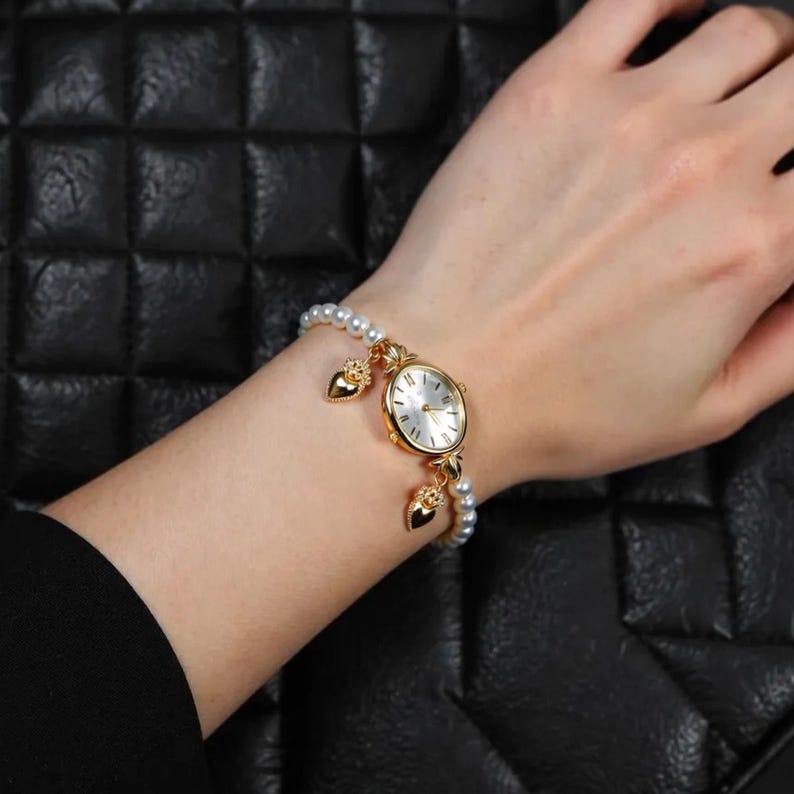 May include: A gold-toned wristwatch with a white face and pearl bracelet. The watch features heart-shaped charms and is worn on a wrist. The background is a black, textured surface.