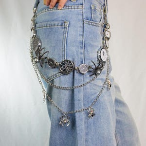 May include: A silver-toned belt featuring multiple watch faces and spider charms. The belt is composed of watch faces, chains, and decorative spiders. The belt is worn over blue jeans.
