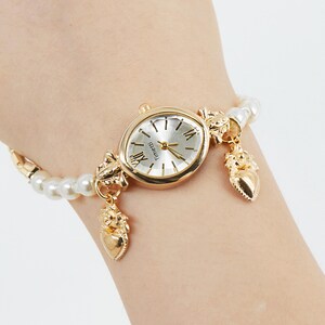 May include: A gold-toned wristwatch with a silver face and Roman numeral hour markers. The watch is attached to a pearl and gold-toned bracelet with decorative charms. The watch face reads "Timeiti".