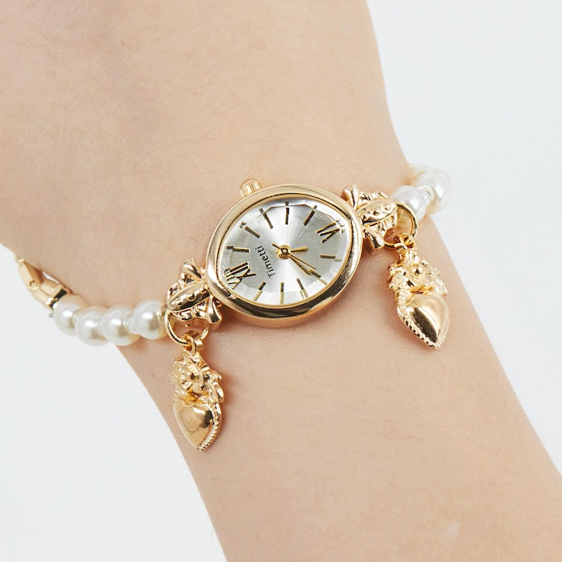 May include: A gold-toned wristwatch with a silver face and Roman numeral hour markers. The watch is attached to a pearl and gold-toned bracelet with decorative charms. The watch face reads "Timeiti".