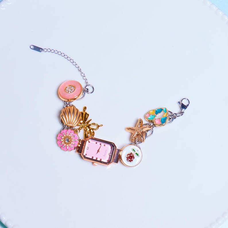 Op de afbeelding: Een gouden armband met bedels, met een roze wijzerplaat en verschillende bedels, waaronder een schelp, een ster, een bloem en een vlinder.