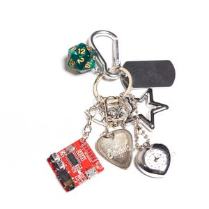 May include: A silver keychain with various charms, including a green and gold 20-sided die, a star, a heart-shaped watch, a heart charm with script, and a red circuit board. The keychain has a silver clasp and a black rectangular tag.