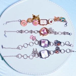 Op de afbeelding: Vier zilveren armbandjes met verschillende wijzerplaten en bedels. De bedels zijn een vlinder, een schelp, een bloem, een hart, een ster en een palmboom.