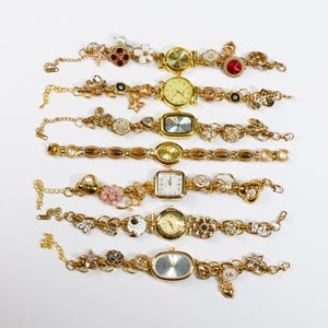 Op de afbeelding: Een verzameling van zeven goudkleurige armbandhorloges. Elk horloge heeft een andere wijzerplaatvorm en is versierd met decoratieve bedels, waaronder bloemen, harten en edelstenen. De wijzerplaten van de horloges hebben verschillende kleuren en stijlen.