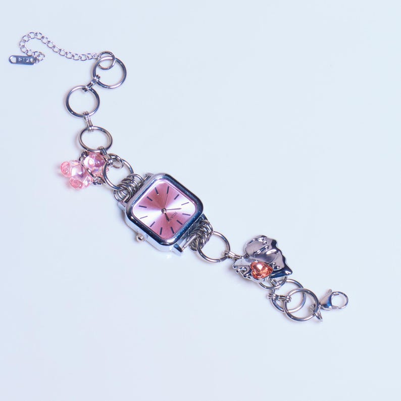 Op de afbeelding: Een zilveren armbandhorloge met een roze wijzerplaat. Het horloge heeft verschillende bedels, waaronder roze en oranje kralen, een zilveren hartbedel en een zilveren sleutelbedel.