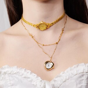 May include: A gold-toned watch choker and a layered necklace with a heart-shaped pendant. The watch choker features a rectangular watch face. The necklace has a delicate chain with small gold beads and a heart-shaped watch pendant.