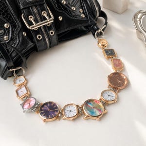 May include: A black textured handbag with silver-tone hardware and a decorative strap composed of multiple connected analogue wristwatches. The watch faces vary in shape, size, and colour, including gold, silver, white, pink, purple, and multi-coloured dials.