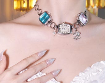May include: A silver-toned watch necklace featuring three watch faces in teal, pink, and white. The necklace includes a pearl and a rose charm. The watch faces have Roman numeral hour markers and hands.