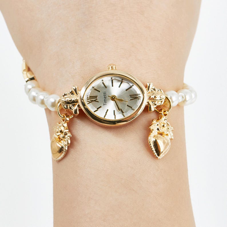 May include: A gold-toned wristwatch with a silver face and Roman numeral hour markers. The watch is attached to a pearl bracelet. The watch face reads "Timetti". Two gold charms hang from the watch. The watch is on a wrist.
