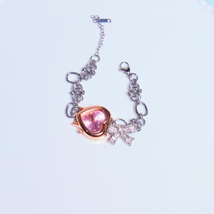 Op de afbeelding: Een zilveren kettingarmband met een roze hartvormig horloge en een strikbedel met strass.