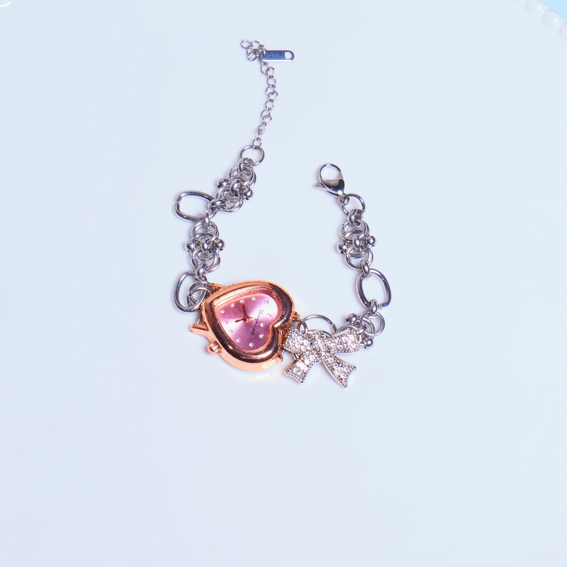Op de afbeelding: Een zilveren kettingarmband met een roze hartvormig horloge en een strikbedel met strass.