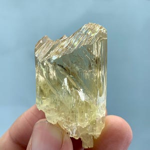 Könnte beinhalten: Ein durchscheinendes, blassgelbes Kristall-Exemplar mit einer prismatischen, länglichen Struktur. Das Mineral weist ausgeprägte vertikale Streifen, scharfe Kanten und eine glasartige, reflektierende Oberfläche auf, die das Licht einfängt und interne Brüche offenbart.