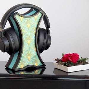 Könnte beinhalten: Ein schwarzer Kopfhörerständer mit einem geometrischen Muster in Teal und Gold. Der Ständer hält ein Paar schwarze Kopfhörer. Der Ständer steht auf einer schwarzen Oberfläche mit einer kleinen weißen Schale mit roten Blumen.