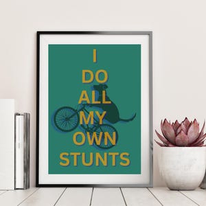 Op de afbeelding: Ingelijste kunstprint met een teal achtergrond, met een hond op een fiets en de gouden tekst "I DO ALL MY OWN STUNTS". Het kunstwerk wordt gepresenteerd op een wit houten oppervlak, met boeken en planten.