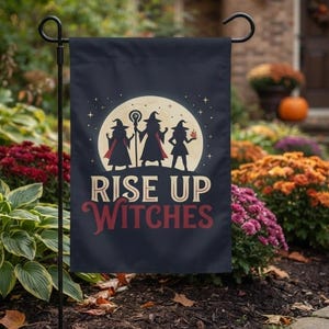 May include: A dark blue garden flag featuring a large yellow full moon silhouette with three witch figures standing in front. The text reads RISE UP in cream and WITCHES in red. The flag is mounted on a black metal stand.