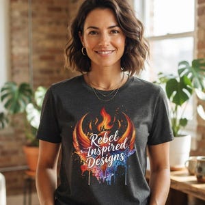 May include: A dark charcoal gray short-sleeve t-shirt featuring a colorful graphic of a stylized phoenix rising from flames. The text Rebel Inspired Designs is printed in white script across the center of the vibrant orange, red, and blue design.