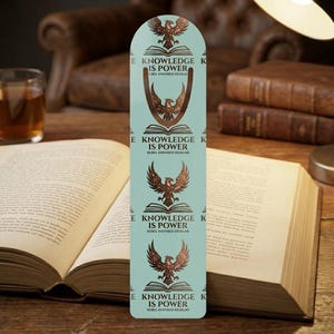 Könnte beinhalten: Ein hellblaues Lesezeichen mit einem sich wiederholenden Muster aus einem offenen Buch mit einem darüber fliegenden Adler, begleitet von dem Text "KNOWLEDGE IS POWER" und "REBEL INSPIRED DESIGNS" in dunkelbrauner Farbe im vertikalen Design.