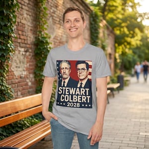 May include: A gray t-shirt featuring a graphic of Stewart and Colbert with the text "STEWART COLBERT 2028" in a navy blue and white design. The shirt is being worn by a person in a casual outdoor setting.