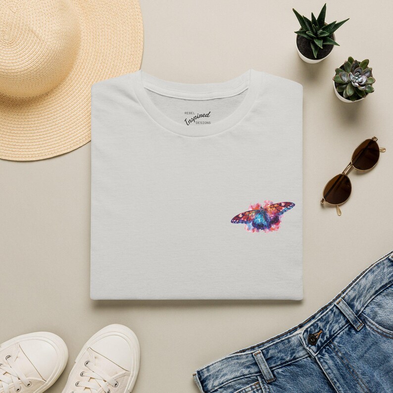 May include: A light gray short-sleeved t-shirt featuring a small, colorful butterfly graphic on the chest. The butterfly design incorporates vibrant shades of blue, pink, and orange. The inner collar displays the text Rebel Inspired Designs.