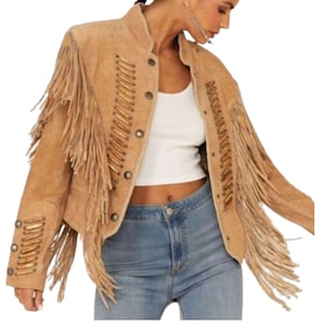 May include: A tan suede jacket with fringe detailing on the sleeves and chest. The jacket has a round collar, button closures, and decorative stitching. The sleeves and back of the jacket are adorned with long fringe.
