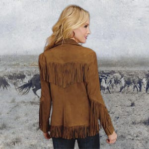 K&ouml;nnte beinhalten: Braune Wildlederjacke mit Fransen am R&uuml;cken, an den &Auml;rmeln und am Saum. Die Jacke hat einen klassischen Western-Stil mit einem Kragen. Der Hintergrund zeigt ein Sepia-Bild einer Rinderherde.