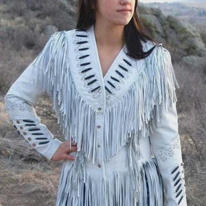 Könnte beinhalten: Weiße Lederjacke mit langen Fransen. Die Jacke hat einen V-Ausschnitt, eine Knopfleiste vorne und dekorative Nähte an Schultern und Ärmeln. Die Ärmel haben schwarze Ausschnitte. Ein stilvolles Kleidungsstück im Western-Stil.