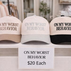 May include: Three baseball caps with the text "ON MY WORST BEHAVIOR" in different color combinations: tan and beige, light gray and beige, and black and beige. The caps are displayed on a white shelf.