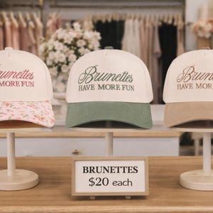 May include: Three baseball caps on display, each with the phrase "Brunettes Have More Fun." One cap has a floral print brim, another a green brim, and the third a tan brim. The caps are displayed on white stands.