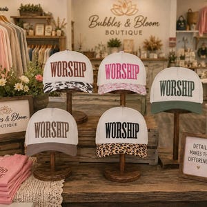 May include: Five baseball caps displayed on wooden stands, each featuring the word WORSHIP embroidered on the front. The caps have white crowns and various brims, including black and white cow print, pink floral, solid olive green, solid gray, and leopard print.