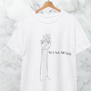 May include: White t-shirt with a minimalist line drawing of a hand making the 'okay' gesture. The text "NI UNA MENOS" is printed below the arm. The shirt is hanging on a wooden hanger.
