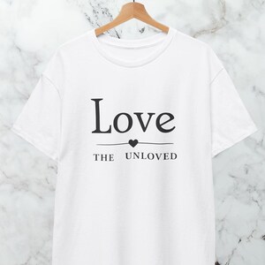 Può includere: T-shirt bianca con le parole "Love" sopra un cuore e una linea, e "THE UNLOVED" sotto. La maglietta è appesa a una gruccia di legno su uno sfondo di marmo.