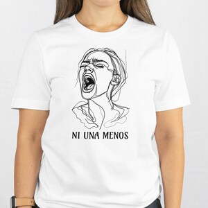 Può includere: T-shirt bianca con un disegno a tratto nero di un volto che urla e la scritta "NI UNA MENOS" sotto. La maglietta è a girocollo. L'immagine è una grafica semplice e audace.