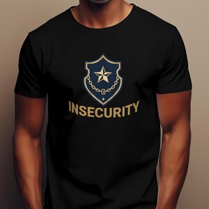 Può includere: T-shirt nera con un design a scudo oro e blu navy, una stella e una catena, e la scritta "INSECURITY" in oro. La maglietta è a girocollo.