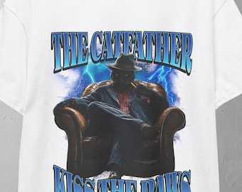 May include: White t-shirt with a graphic of a person in a hat and suit seated in a chair, with the text "THE CATFATHER MISS THE PAWS" in blue. The design has a vintage, retro style.