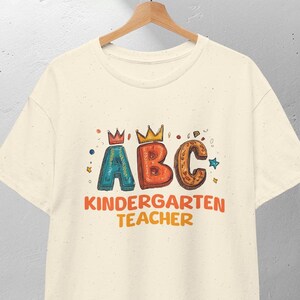 Può includere: T-shirt color crema con la scritta "ABC Kindergarten Teacher" in lettere colorate e giocose. Le lettere A, B e C sono decorate con corone. La maglietta è appesa a una gruccia di legno.
