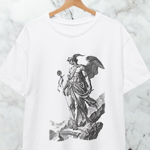 Pode incluir: T-shirt branco com uma ilustração a preto e branco de uma figura clássica alada. A obra de arte retrata uma figura detalhada com um bastão e roupas drapeadas. O design está centrado na t-shirt, destacando os detalhes intrincados.