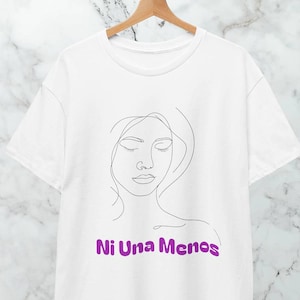 Op de afbeelding: Wit T-shirt met een minimalistische lijntekening van een vrouwengezicht. De tekst "Ni Una Menos" is in paars onder de tekening gedrukt. Het T-shirt hangt aan een houten hanger.