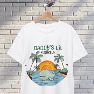 Può includere: T-shirt bianco con la scritta "DADDY'S Lil SQUIRTER" in verde. La maglietta presenta una balena dei cartoni animati nell'oceano con palme, un tramonto arancione e piccole isole. Il disegno è stampato sul davanti.